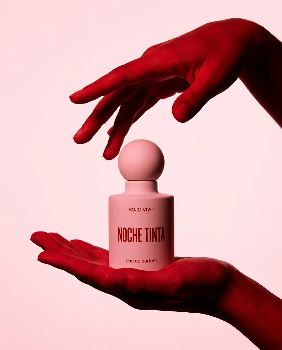 A red hand about to touch the cap of a pink perfume bottle held by another hand, on a pink background.